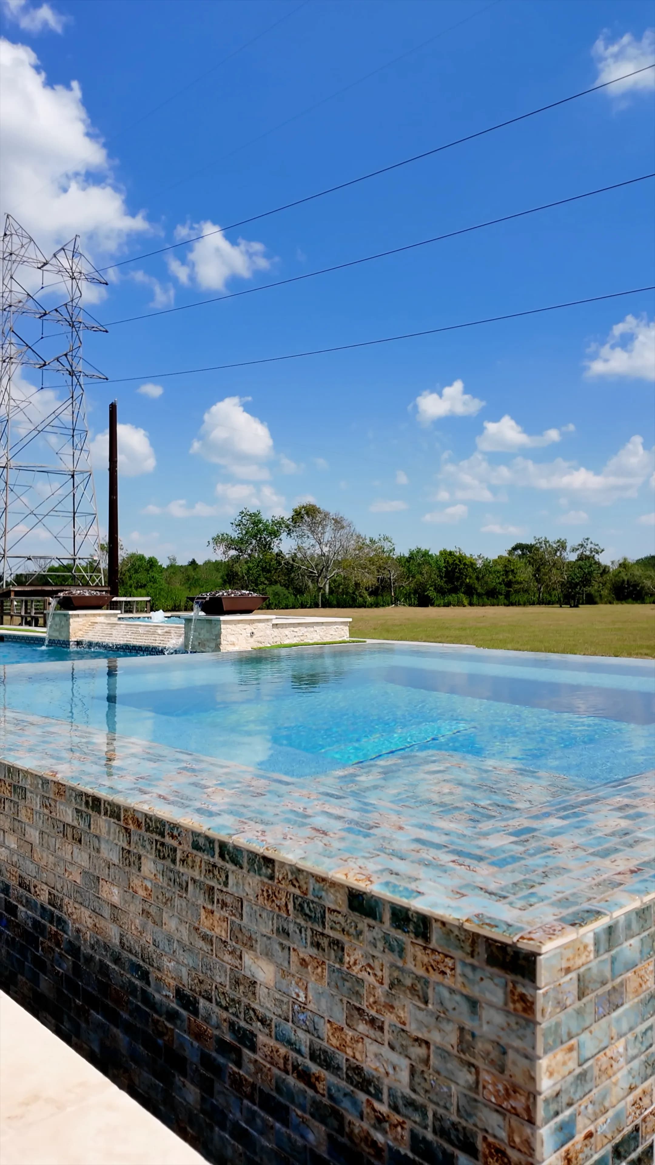 Corner of the perimeter-overflow spa clad in blue-and-amber glass mosaic, with open pasture beyond