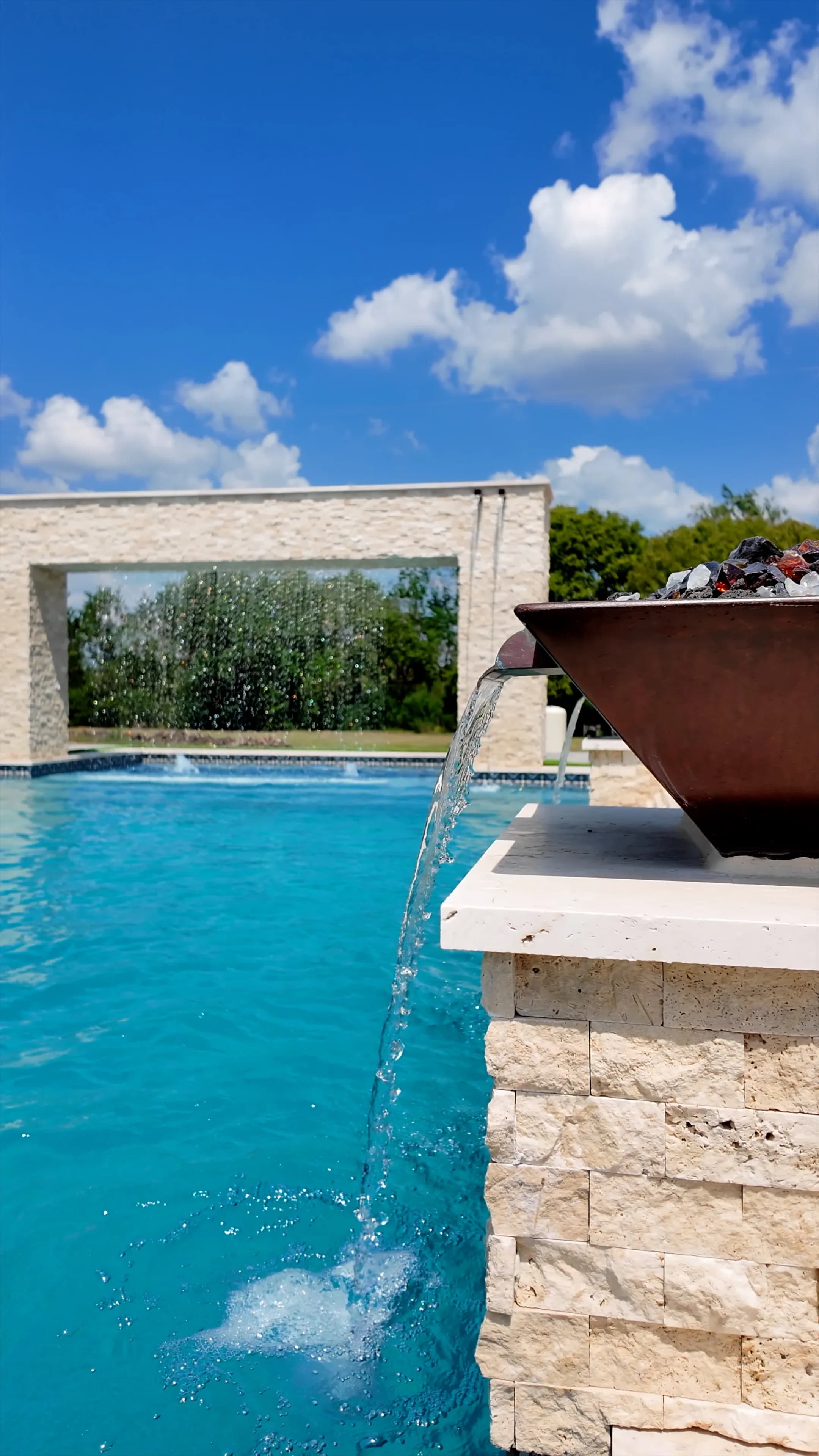 Copper fire bowl spilling water into the pool from its stacked travertine pedestal