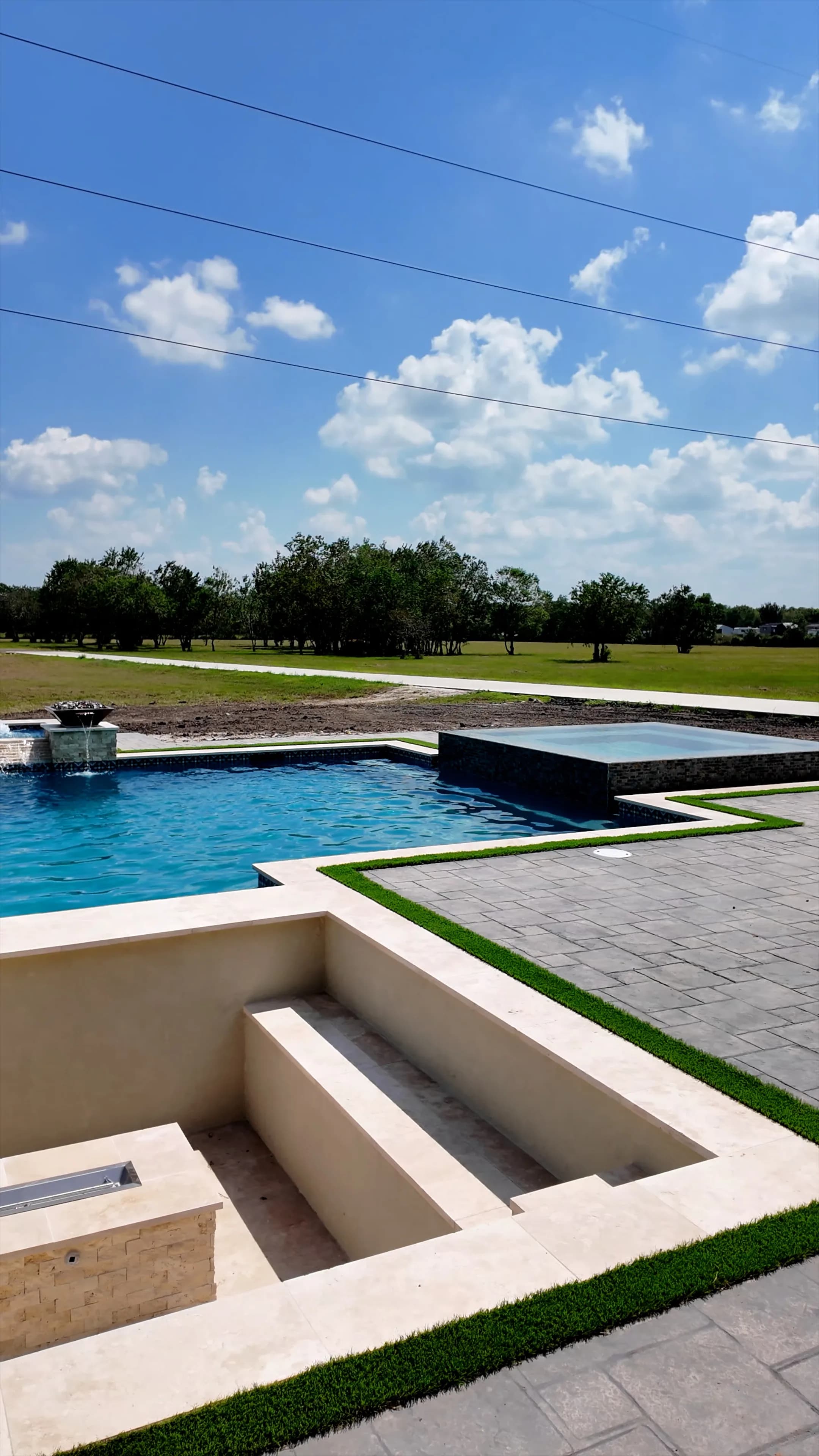 Sunken travertine lounge with linear fire pit beside the pool, raised spa, and open field