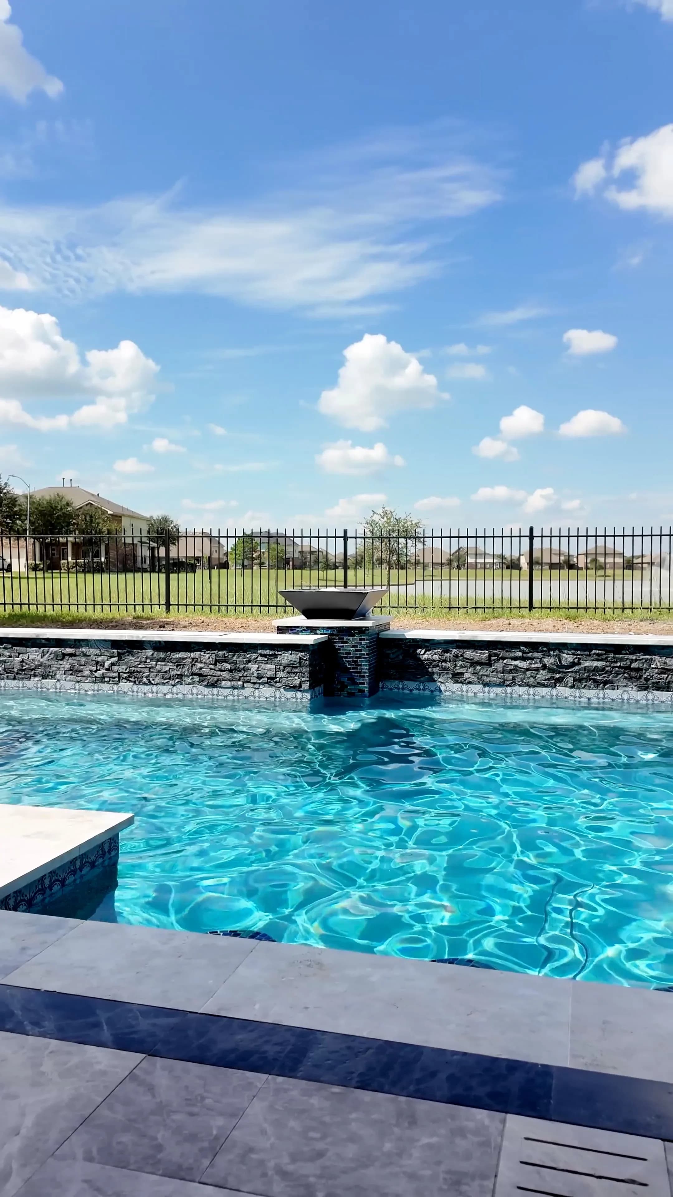 Fire bowl on a glass-tile pedestal above the dark stacked-stone wall, with green space beyond the iron fence
